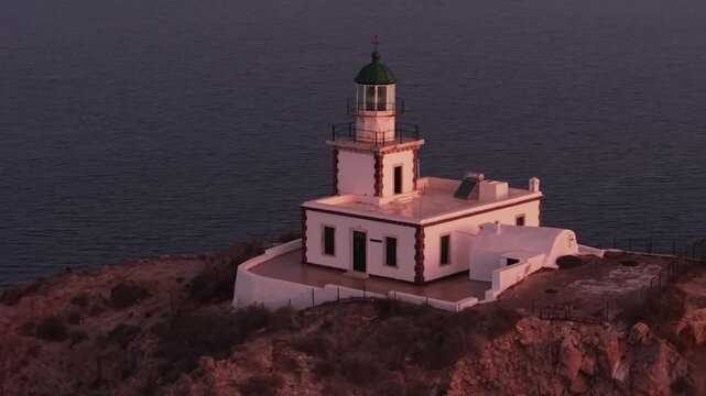 Drone orbit around the Akrotiri Lighthouse in Santorini at sunrise. The camera circles the beacon, bathed in warm golden light against the deep blue Aegean Sea and rugged coastal cliffs.