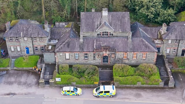 A slow rightward drone pan reveals the full facade of Windermere police station, a traditional Cumbrian slate building, with two liveried police vehicles parked outside on the main road.