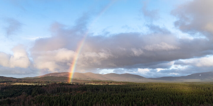 Aerial view of Cairngorms National Park with a bright rainbow arching over dense evergreen forests and rolling mountains under a cloudy sky in Scotland, United Kingdom.