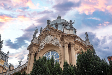 Naklejka premium The ornate Baroque facade of Vajdahunyad Castle in Budapest features grand statues and a prominent dome set against a vibrant pink sunset sky.