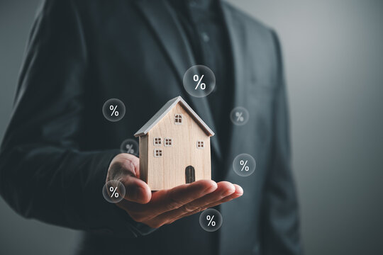 Businessman holding a wooden house with percentage icons, symbolizing mortgage rates, home loans, refinancing, real estate investment, housing finance, and property market decisions.