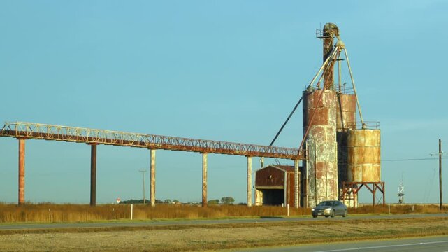 Cactus, Texas - November 06, 2025: a blue Freightliner Cascadia semi-truck traveling on a road past a rural grain storage elevator facility.