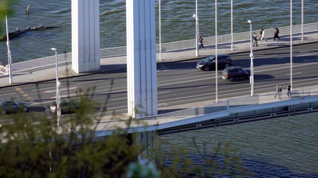 Urban life on a Budapest bridge over the Danube, with pedestrians, traffic and sunlight reflecting on the river in a scenic city view.