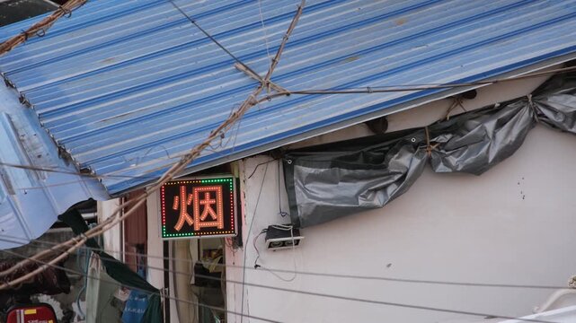 A small Chinese retail shop displaying a glowing LED sign with the Chinese character 烟 (tobacco or cigarettes). Gritty, authentic aesthetic street life in a&nbsp;low-income and run-down urban neighbourhood