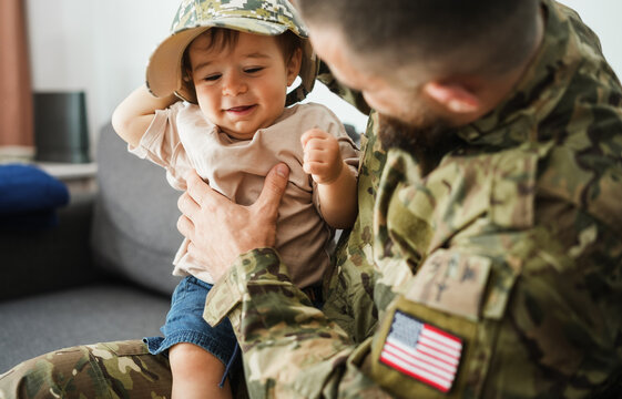 Male american soldier in military uniform hugs his little son at home while baby boy playing with his hat - Emotional happy homecoming concept - Main focus on man face