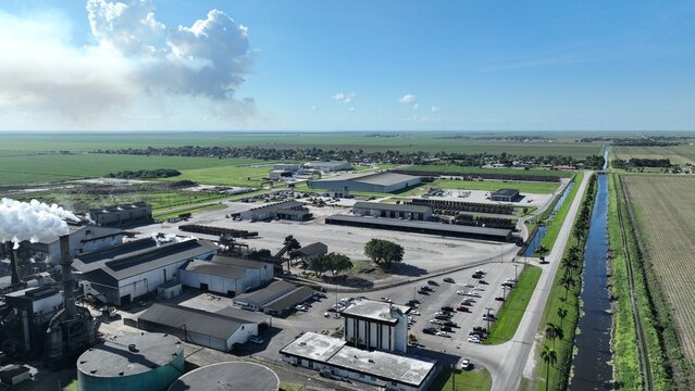 Aerial view of an industrial processing plant with smoke rising from chimneys, large warehouses, and a parking lot next to a canal, palm trees, and green fields.