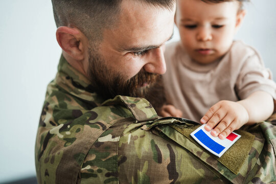 Male french soldier in military uniform hugs his little son at home while baby boy touching french flag - Emotional military happy homecoming concept - Main focus on man eye