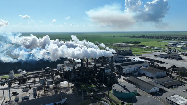 Aerial view of an industrial sugar mill complex with thick white smoke billowing from multiple chimneys over green agricultural fields under a clear blue sky.