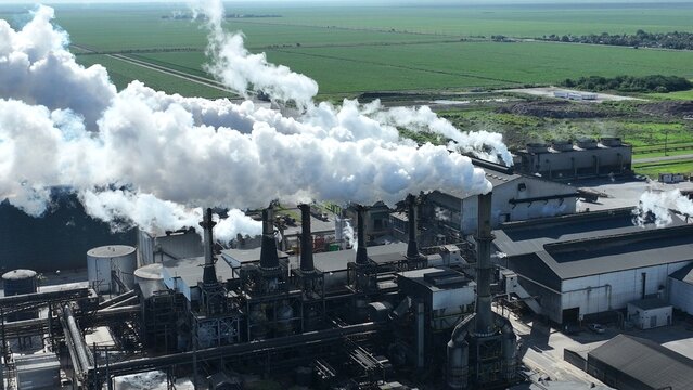 Aerial view of an industrial factory with multiple smokestacks emitting thick white smoke plumes over vast green agricultural fields under a clear sky.