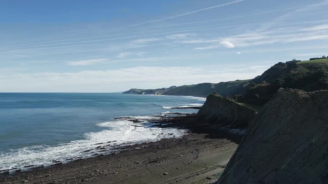Majestic coastal landscape of the Basque Coast Geopark featuring the famous flysch formations of Sakoneta beach in Zumaia, with waves crashing against the rocky shoreline under a clear blue sky