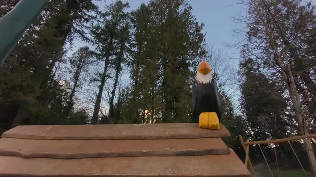 Carved bald eagle statue featuring yellow beak resting on wooden play structure roof framed against tall evergreen trees beneath cloudy dusk sky captured via low angle perspective