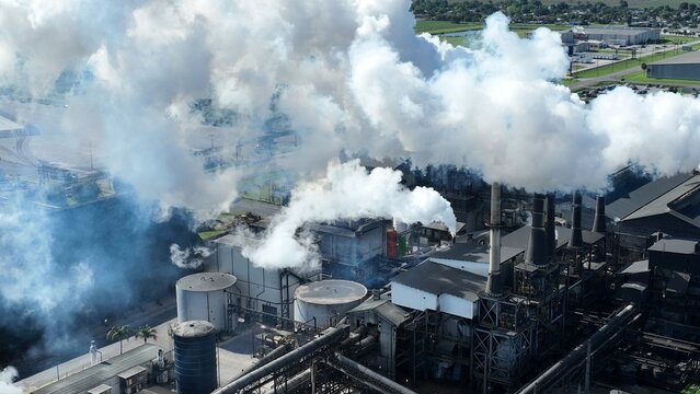 Aerial view of an industrial factory complex with thick white smoke billowing from chimneys and storage tanks.