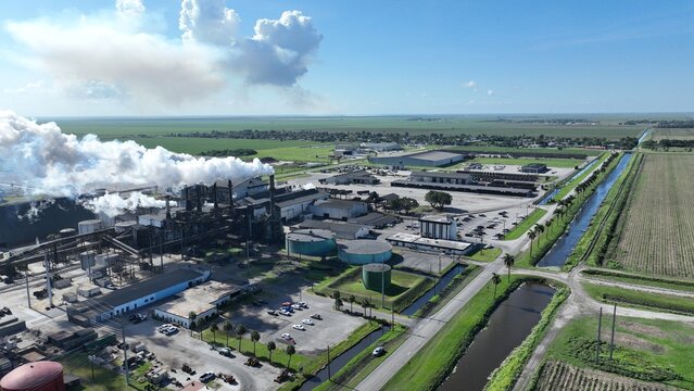 Aerial view of a large industrial facility with smoke rising from chimneys, storage tanks, and surrounding green agricultural fields under a blue sky.