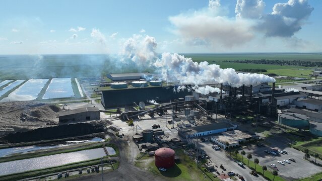 Aerial view of an industrial sugar mill facility with smoke rising from tall chimneys, large storage tanks, and surrounding agricultural fields under a blue sky with white clouds.