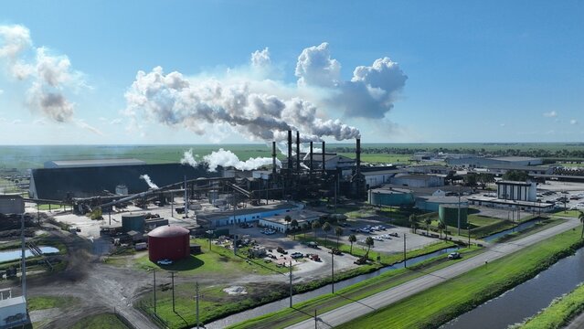 Aerial view of a large industrial facility with multiple smokestacks emitting thick white steam over a flat green landscape with storage tanks and a canal.