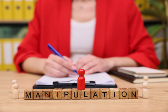 In a vibrant office, a focused individual in red writes notes while a prominent block of letters spells MANIPULATION on the table, surrounded by wooden figures, suggestive of thoughtful analysis.