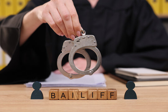 In a serious courtroom setting, a bailiff holds up gleaming handcuffs while two figures watch. Papers and files create a backdrop for this dramatic legal moment.