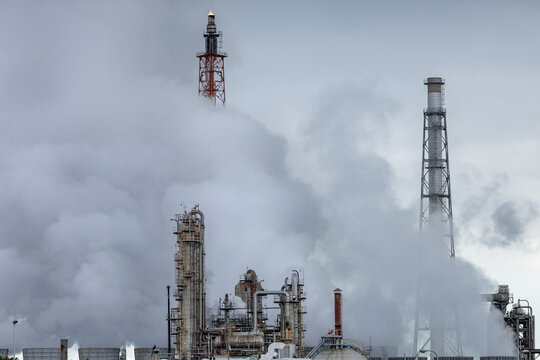 Dense white smoke and steam shroud an industrial refinery's tall distillation and processing tower, with top of a lit flare stack emerging from among the thick, billowing plumes.