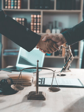 The lawyer is shaking hands with his client to congratulate them on the successful settlement of a legal case, with a gavel and legal scales beside them.