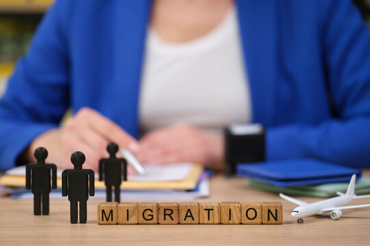 On a wooden desk, three black figures stand beside blocks spelling migration. A woman in a blue jacket is engaged in paperwork, symbolizing a discussion about relocation and movement.