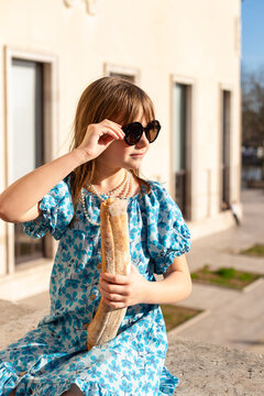 A young girl in a blue floral dress and sunglasses sitting outdoors in Paris while holding a French baguette. She is wearing a pearl necklace and looking to the side.