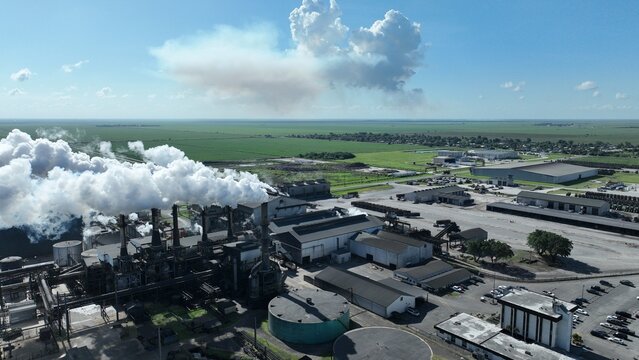 Aerial view of an industrial factory with thick white smoke rising from chimneys near green agricultural fields and storage tanks under a blue sky.