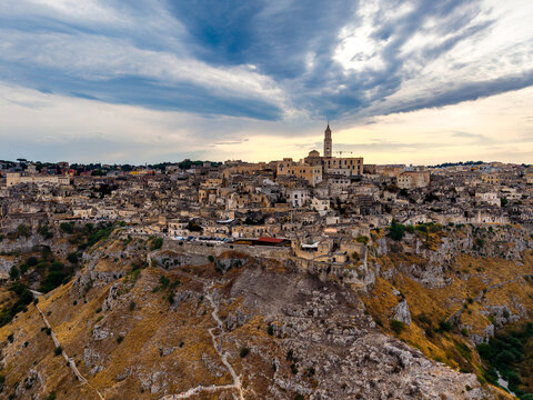 Aerial view of the ancient Sassi di Matera district with its historic stone dwellings and cathedral tower under a dramatic cloudy sky at sunset in Matera, Basilicata, Italy.