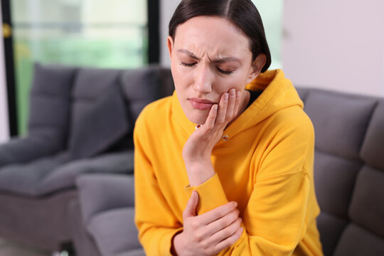 A woman experiences discomfort and pain as she holds her jaw with one hand while seated comfortably on a cozy couch. Soft sunlight pours in, adding warmth to the scene.