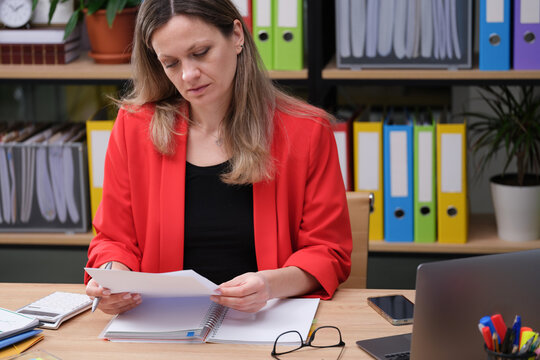 Sitting at a stylish wooden desk, a woman in a red blazer carefully examines important papers. Surrounding her are vibrant binders and a sleek laptop, creating a focused atmosphere.