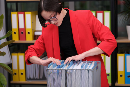 A focused professional in a bright red blazer carefully sorts through files in a stylish, modern office. Bright folders line the shelves, creating a vibrant atmosphere.