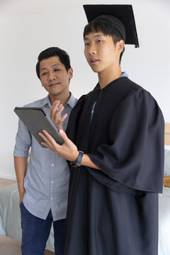 Asian males standing in bedroom, graduate wearing gown and mortarboard holding tablet, gesturing