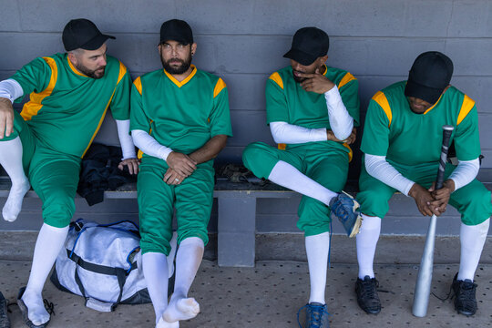 Diverse male teammates sitting on dugout bench wearing green jerseys holding bat and duffel bag