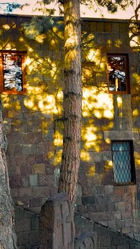 Ancient Stone Cross Khachkar by Stone Building in Armenia. Historic carved stone khachkar monument nestled between pine trees and a stone building for cultural travel and heritage promotion.