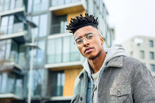 A young man with glasses and styled hair stands confidently in front of a modern apartment building.