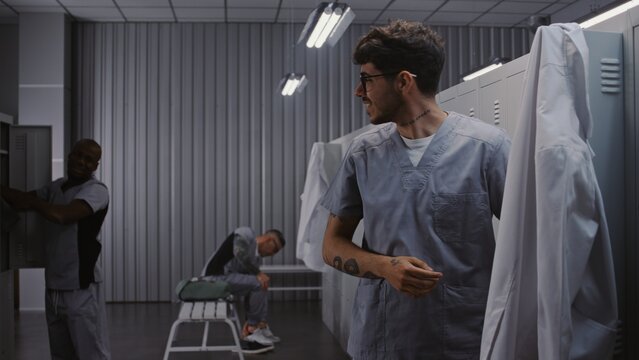 Diverse Group of Male Doctors in Surgical Scrubs and Lab Coats Interact by Grey Lockers, Hanging Up Uniforms and Preparing to Leave Hospital After Shift. Concept of Professional Medical Environment.
