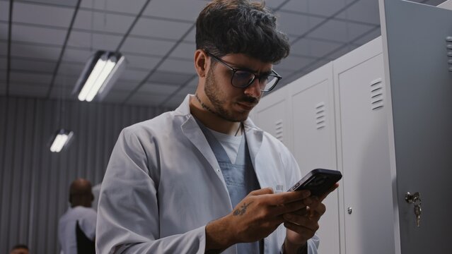 Young Male Physician in White Coat Stands by Locker After Shift, Intensely Reviewing Urgent Laboratory Results and Patient Diagnostics on Smartphone. Concept of Medical Urgency and Dedication.