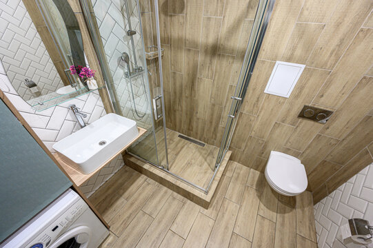 A compact modern bathroom with wooden-textured tiles, glass shower cabin, wall-mounted sink, minimalist toilet, washing machine, herringbone white tiles, glass shelf with flowers, chrome fixtures.