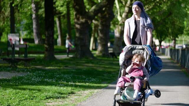 Mother with headscarf pushing toddler in stroller along sunny park path, cancer recovery and chemotherapy journey, hope and resilience during difficult times in family life