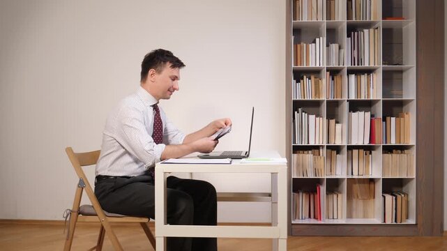 Businessman sitting at a desk, looking at a laptop screen and happily counting earned money in an office environment with a bookshelf in the background