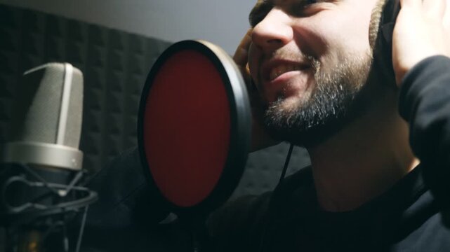 Male singer in headphones singing song into the microphone at sound studio. Young man emotionally recording new melody or song. Working of creative musician. Show business concept. Slow motion