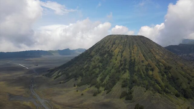 4K aerial view of Mount Batok&rsquo;s green slopes and the vast Bromo volcanic sand sea under blue skies.
