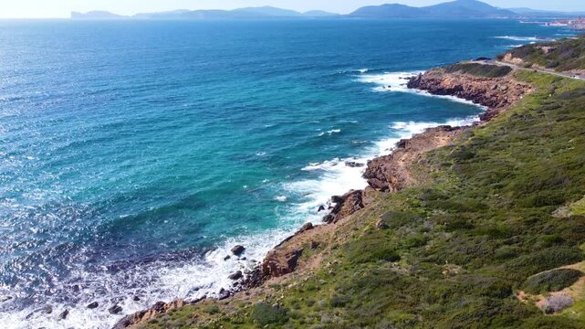 Aerial drone rising shot over the southern coast of Alghero with Capo Caccia visible on the horizon, clear sea and rugged Mediterranean landscape