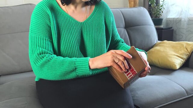 Woman in green sweater opens a gift box on a gray couch, revealing contents while sitting in a cozy living room with plants and natural light