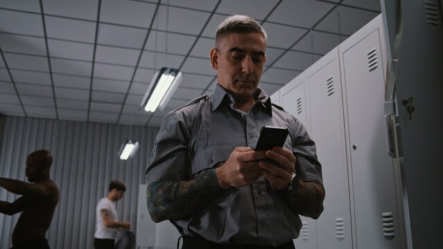 Male Security Officer With Arm Tattoos Hangs ID Badge on Locker Door, Takes Out Smartphone, and Checks For Missed Calls and Messages After Finishing Workday. Shift End Concept in Staff Dressing Room.