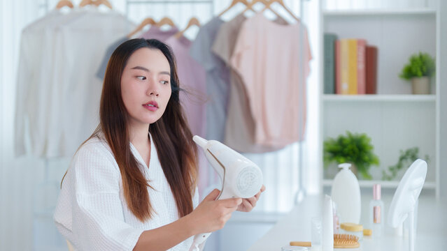 Asian woman using hair dryer blow drying hair beauty morning routine grooming standing in modern bedroom closet room with clothing rack mirror getting ready
