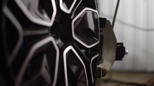 Macro close-up of a glossy black alloy wheel with intricate geometric spokes beside a mounted brake caliper in an automotive workshop, highlighting precision machining and finishes