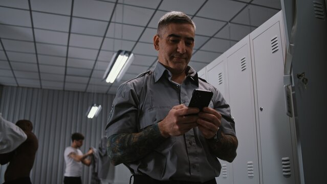 Male Security Officer With Arm Tattoos Hangs ID Badge on Locker Door, Takes Out Smartphone, and Checks For Missed Calls and Messages After Finishing Workday. Shift End Concept in Staff Dressing Room.