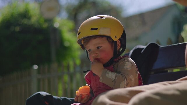 Toddler in yellow helmet sitting on bench outdoors, tasting tangerine segment then pausing to observe it closely, curious sensory moment exploring flavor and texture in sunlight