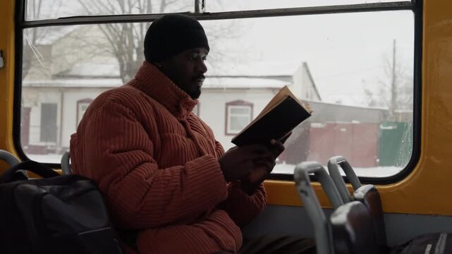Black commuter reading book on bus frames portray student, writer, traveler winter suburban houses pass by orange puffer jacket and black beanie backpack on adjacent seat soft natural light