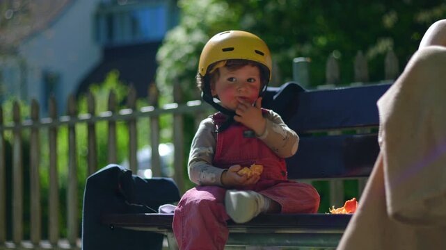 Toddler wearing yellow helmet sitting on park bench, eating tangerine segments in warm sunlight, candid outdoor moment of childhood curiosity and simple everyday snacking